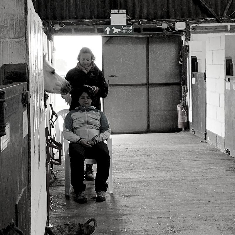 Black and white picture of Ali in a stable giving reiki to a lady sat on a chair