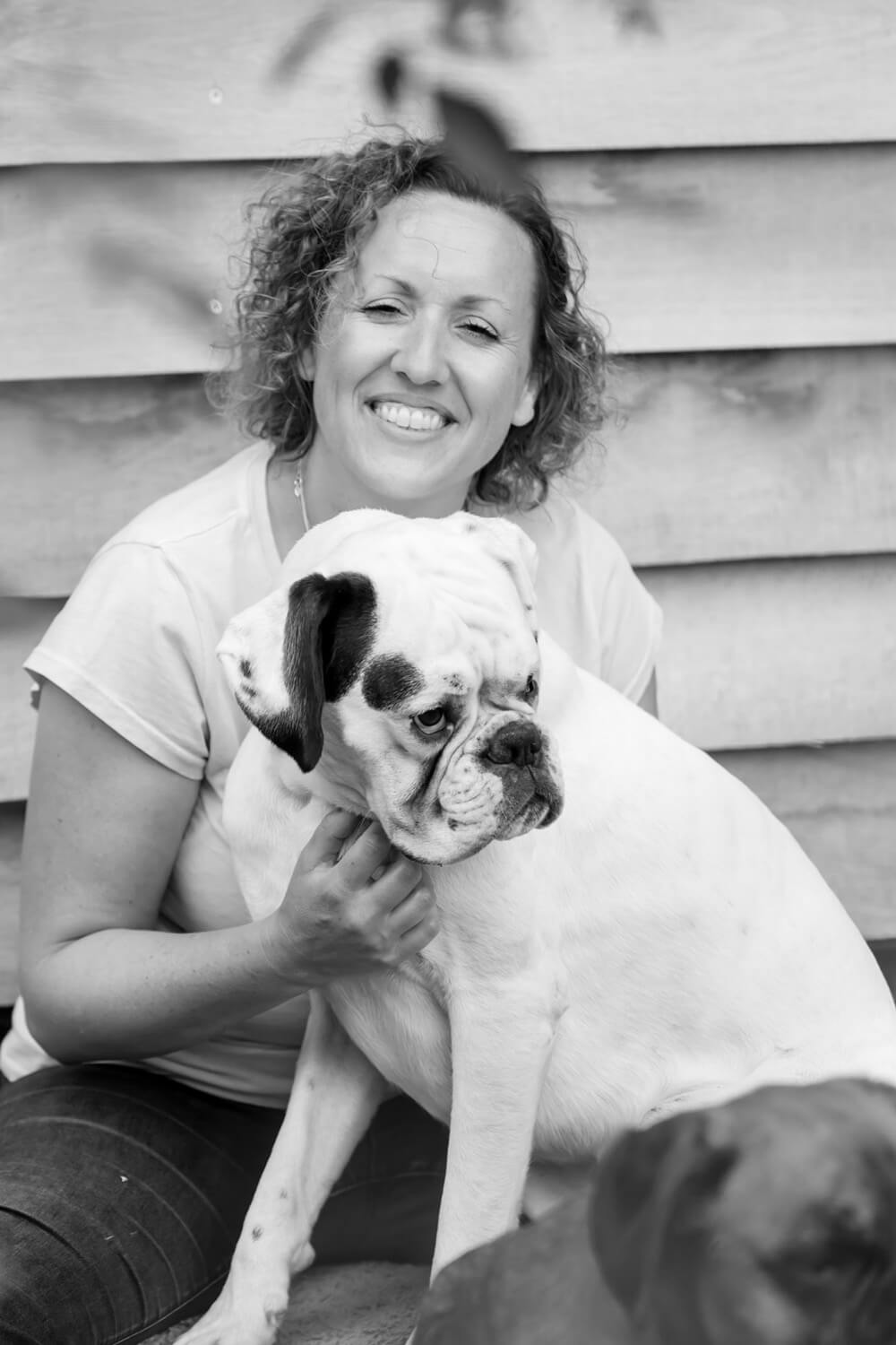 Ali Carrington a Dog Listnener, Dog Behaviour Specialist and Animal Reiki Master sitting, smiling, facing the camera with her hand gently on her dog a white Boer called Ludo who looks to the right hand side